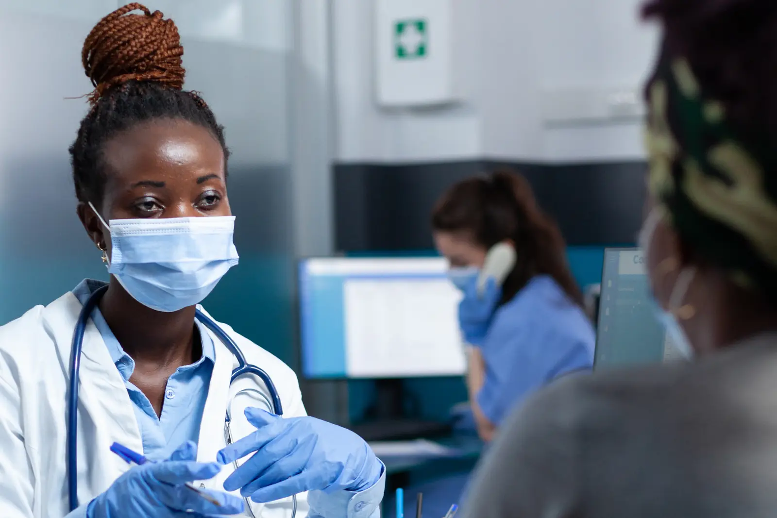 A clinician in a mask and gloves talking with a patient across a desk in a clinic, with a workstation behind her