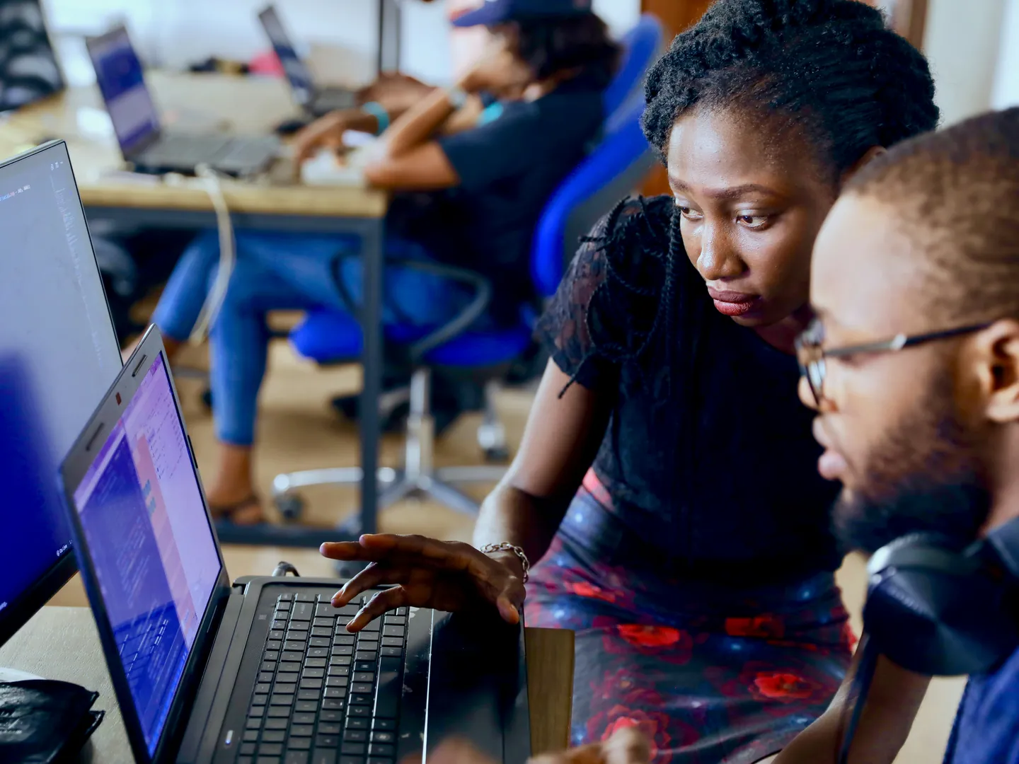 Two developers working through code together on a laptop in a busy software studio